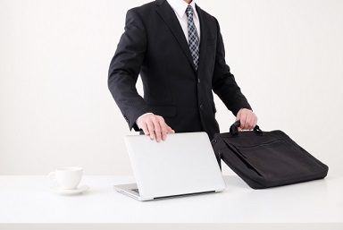 スーツ姿の男性がデスクの上でノートパソコンとビジネスバッグを扱っている様子